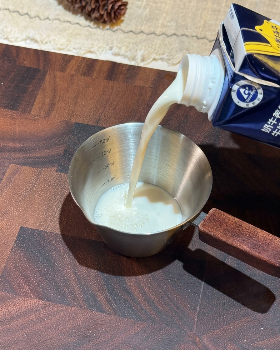 Weiße Milch wird aus einem blauen Karton in einen kleinen Edelstahl-Messbecher mit Holzgriff gegossen.