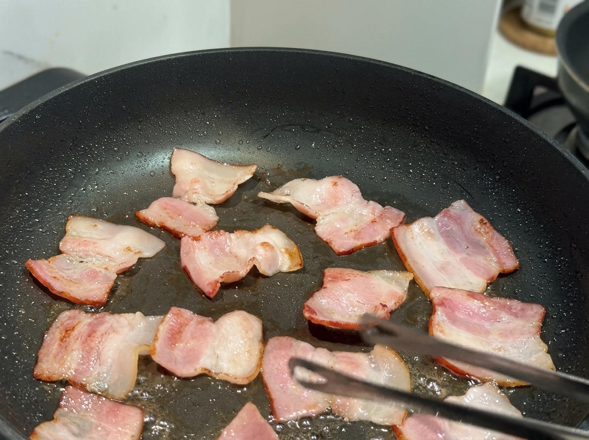 Kleine, flache Baconstücke werden in einer schwarzen Antihaft-Pfanne gebraten. Eine Küchenzange ist sichtbar.