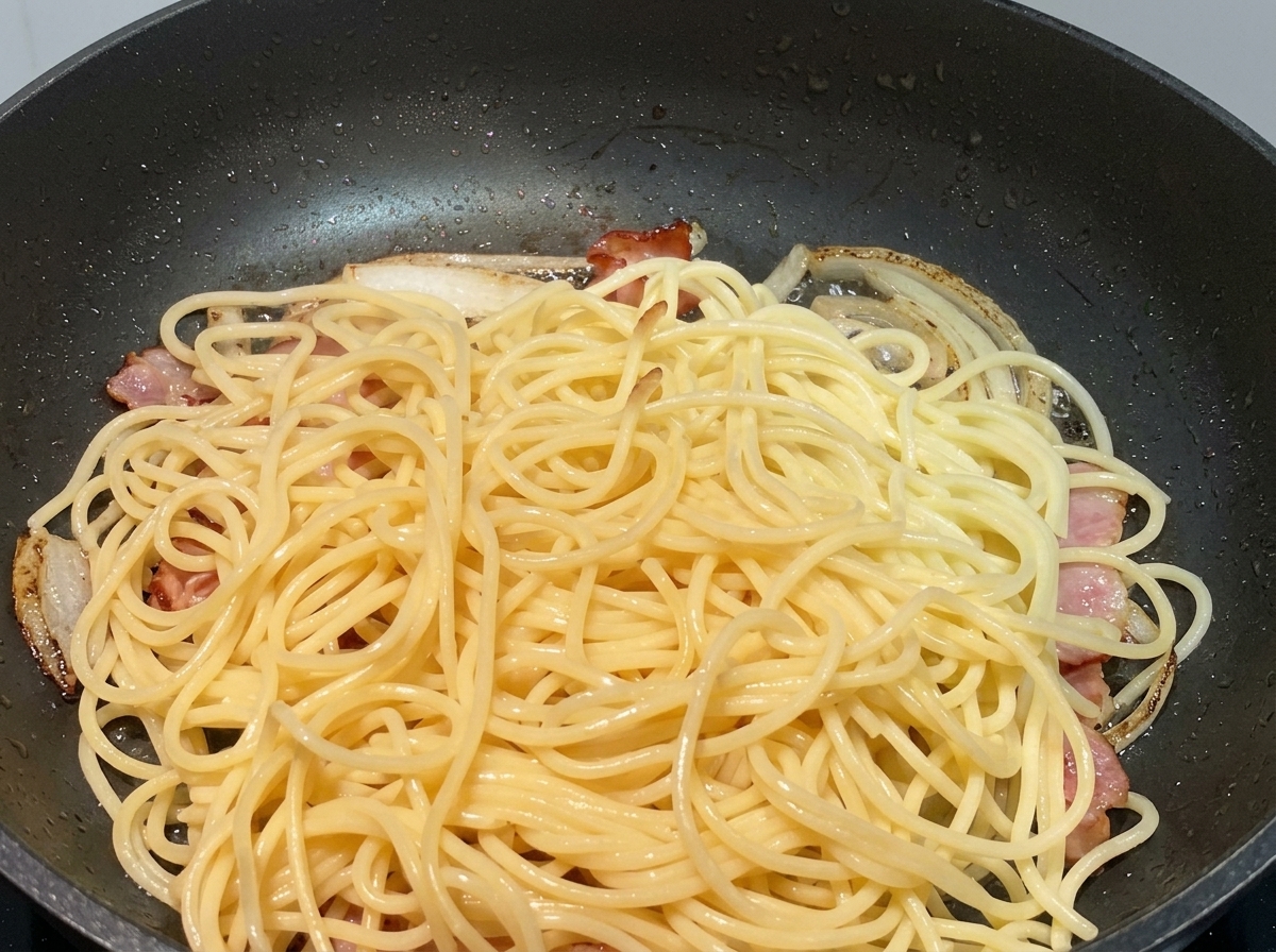 Gekochte Spaghetti-Nudeln liegen auf angebratenen Zwiebeln und Baconstücken in einer schwarzen Pfanne.