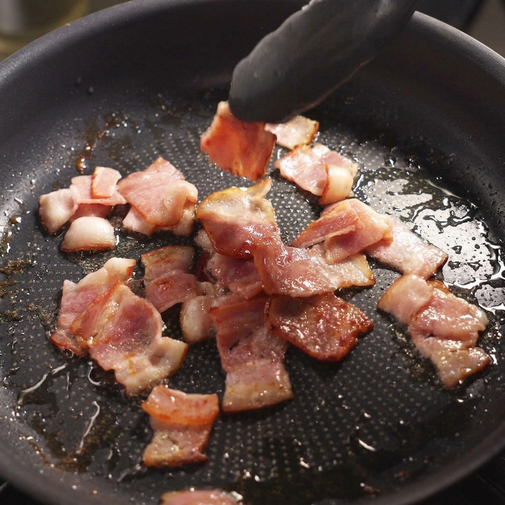 Eine schwarze Zange wendet leicht gebräunten, brutzelnden Speck in einer Pfanne.