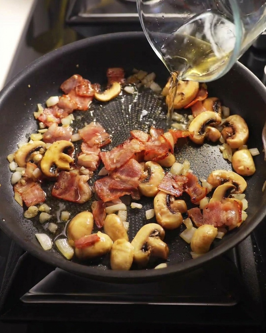 Klarer Weißwein wird aus einem Messbecher in eine Pfanne mit Pilzen und Speck gegossen.