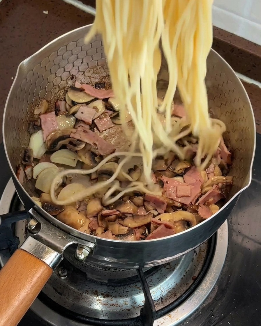 Gekochte Spaghetti werden in eine Pfanne mit angebratenen Pilzen und Speck gegeben.