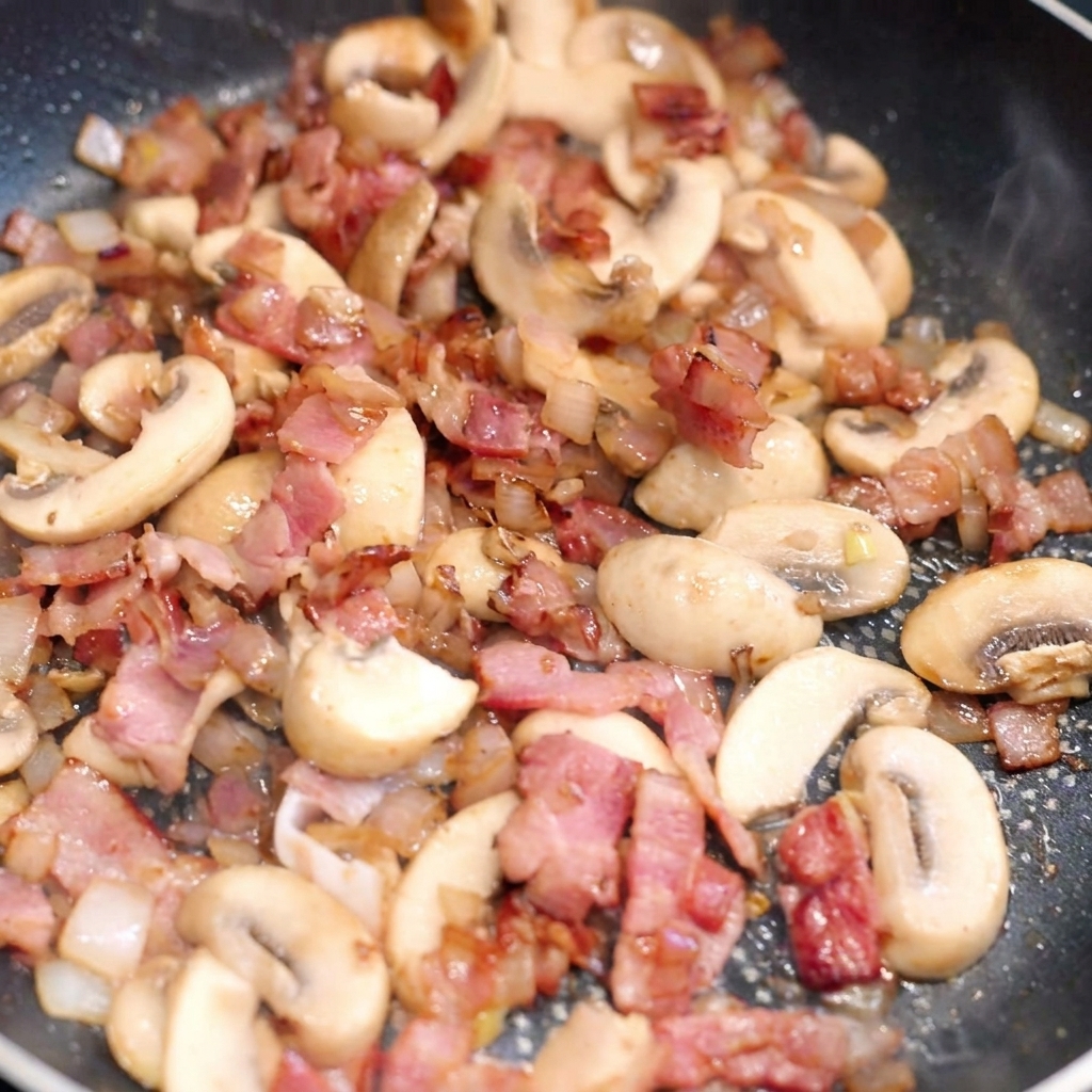 Breite Nudeln werden in einer Pfanne mit gebräuntem Speck, sautierten Pilzen und Zwiebeln vermengt.