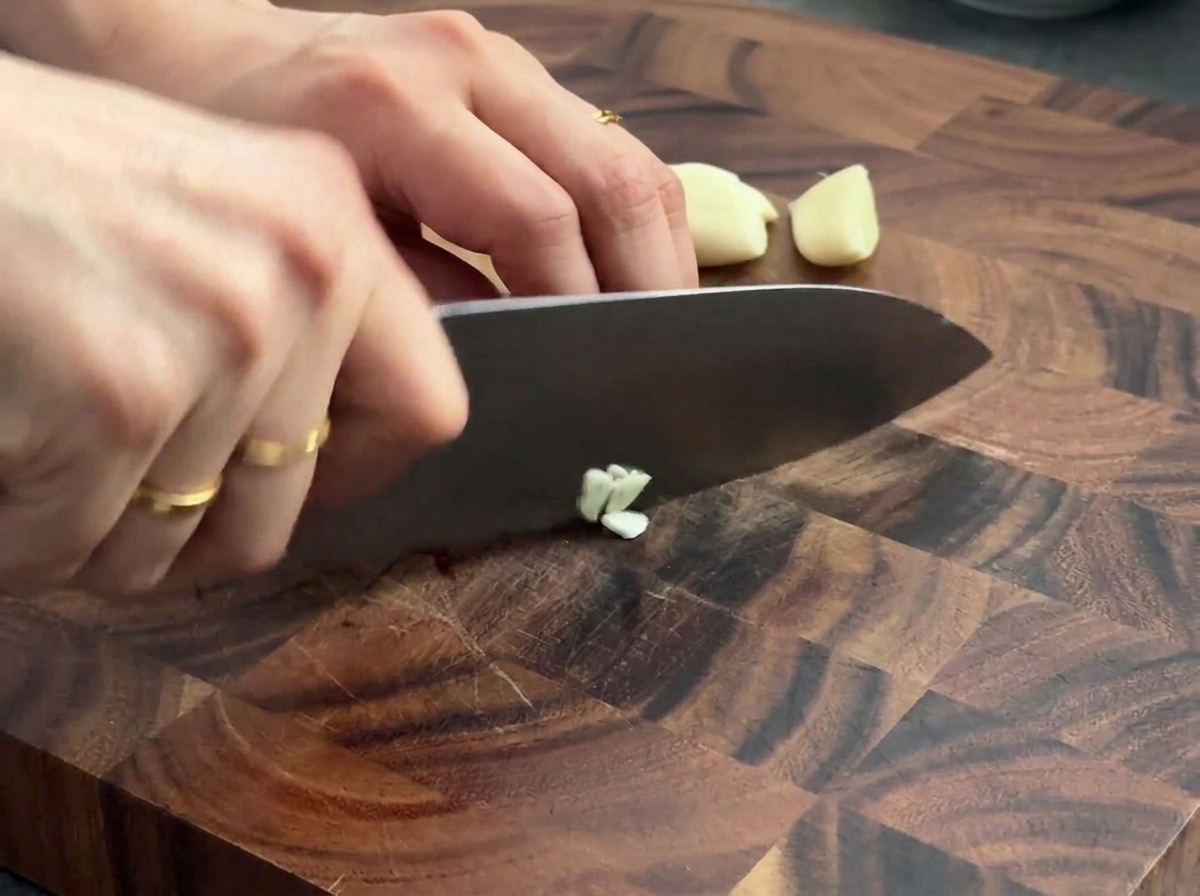 Frische Knoblauchzehen werden auf einem Holzschneidebrett mit einem Kochmesser zerkleinert.