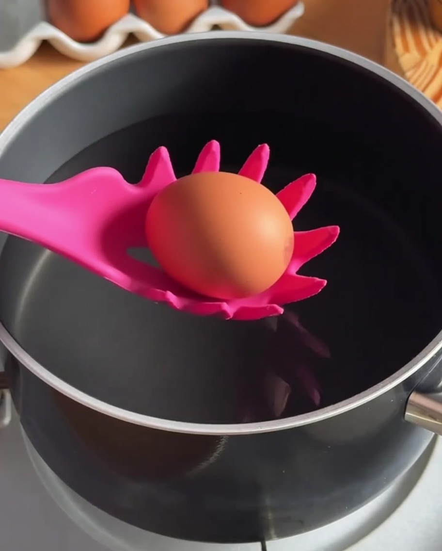 Ein Ei wird mit einem Schaumlöffel aus Silikon in heißes Wasser gegeben.