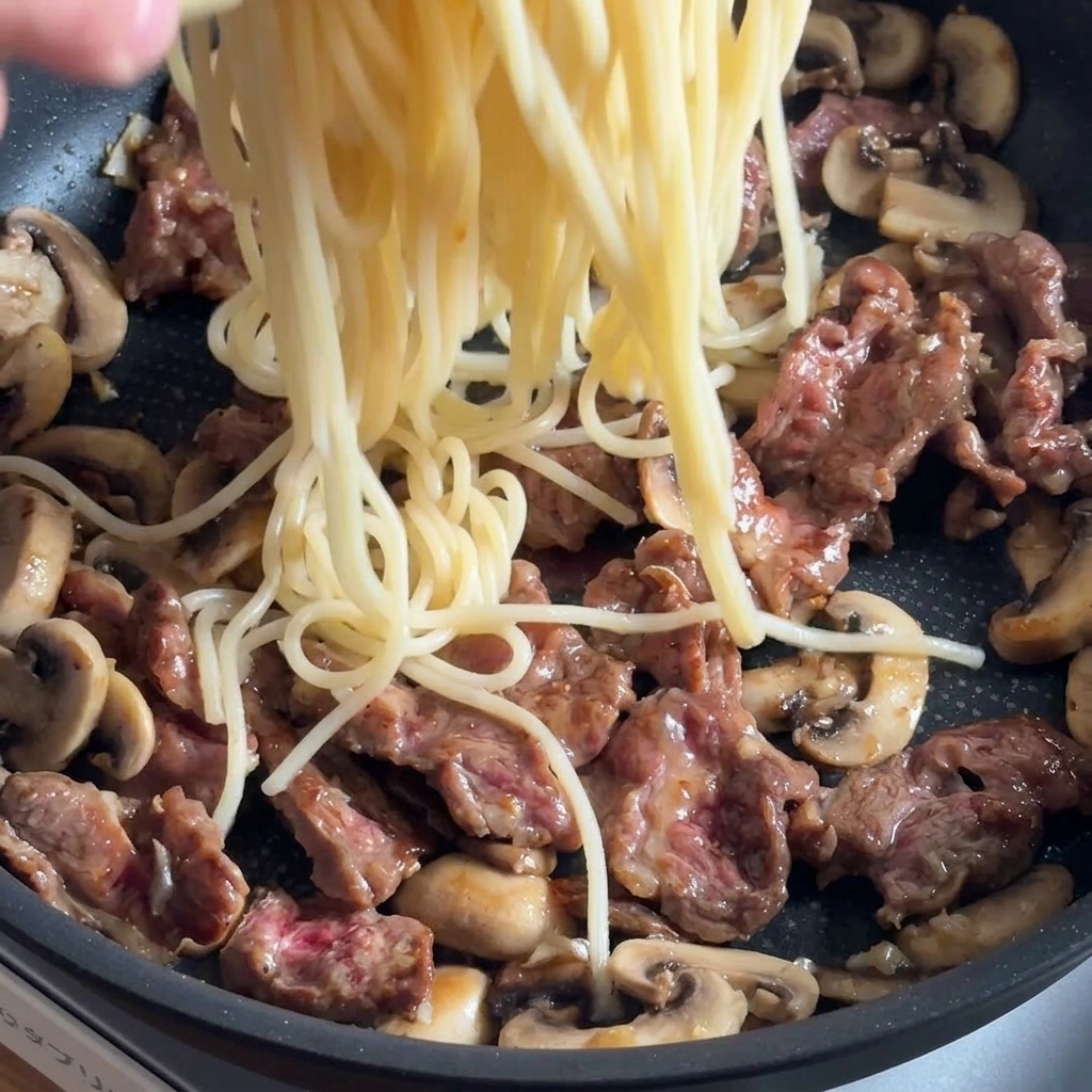 Pasta wird in die Pfanne zu Rindfleisch und Pilzen gegeben, um mit den Saucen vermischt zu werden.