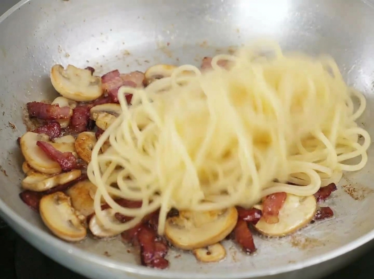 Spaghetti werden in einer Pfanne mit gebratenem Speck und Pilzen geschwenkt.
