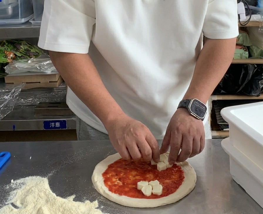 Hände legen frische Mozzarellawürfel auf einen mit Tomatensauce bestrichenen Pizzateig.