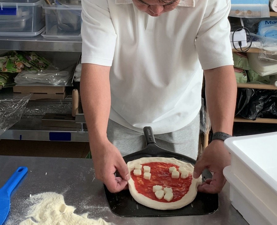 Hände justieren die runden Ränder einer ungebackenen Margherita-Pizza auf einem gelochten Pizzaschieber aus Metall.