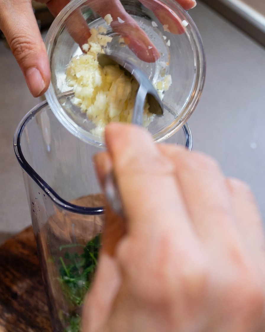 Ein Mixbehälter wird mit gehacktem Knoblauch, frisch geriebenem Käse und gerösteten Pinienkernen gefüllt.