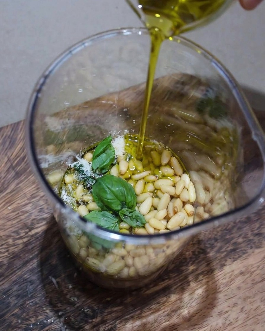 Nahaufnahme von Pesto-Zutaten, die im Behälter gemixt werden, während ein dünner Strahl Olivenöl hinzugefügt wird.