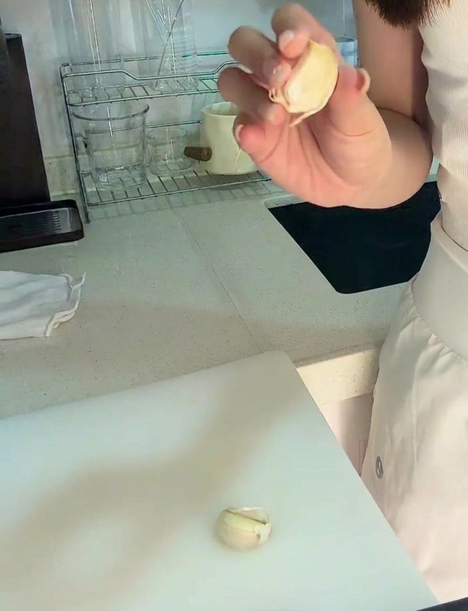 Eine Hand hält eine geschälte Knoblauchzehe über ein weißes Schneidebrett, auf dem bereits etwas gehackter Knoblauch liegt.
