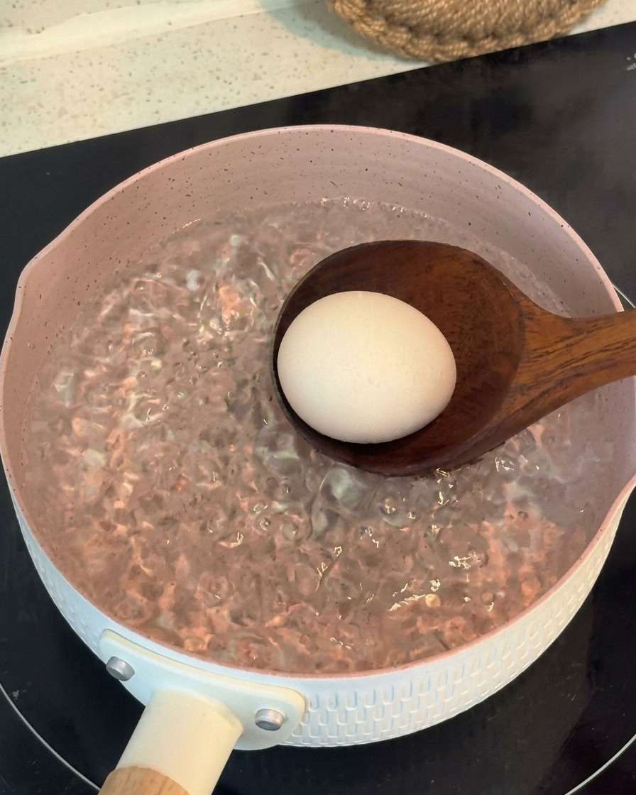 Eine Schöpfkelle lässt ein ganzes Ei vorsichtig in einen rosa Topf mit sprudelnd kochendem Wasser gleiten.