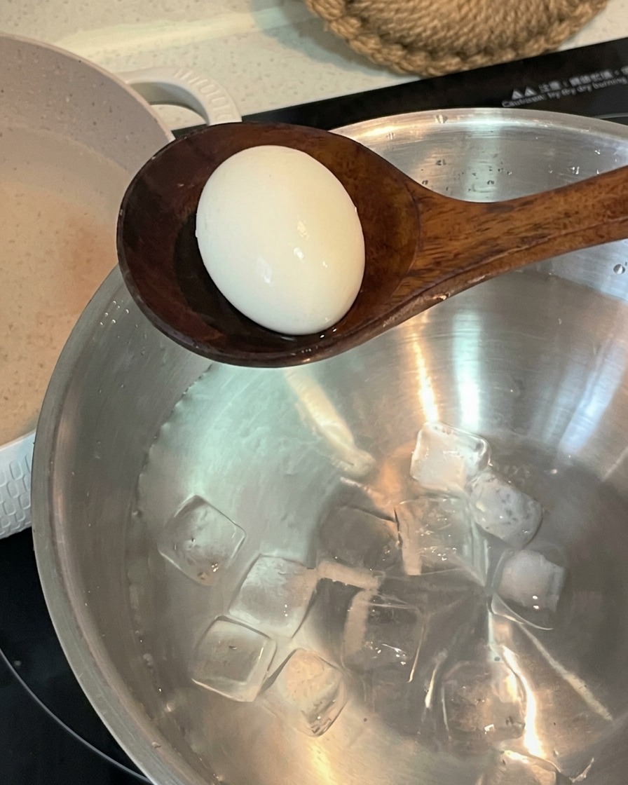 Ein Holzlöffel befördert ein gekochtes Ei in eine Edelstahlschüssel mit kaltem Wasser und Eiswürfeln.