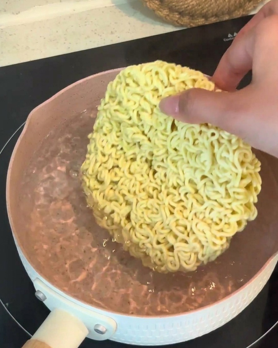 Eine Hand legt einen trockenen, runden Block Instant-Ramen-Nudeln in einen Topf mit kochendem Wasser.