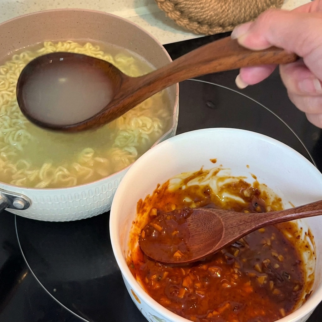Heißes Nudelwasser wird aus einer Holzschöpfkelle in eine Schüssel mit der gemischten Gewürzpaste gegossen.