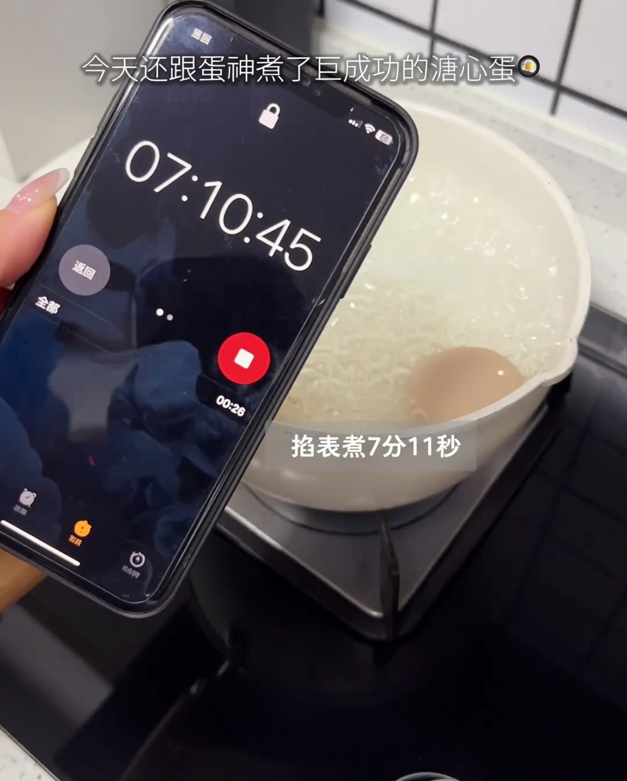 Ein rohes Ei kocht in einem Topf auf dem Herd, im Vordergrund ein Smartphone-Timer.