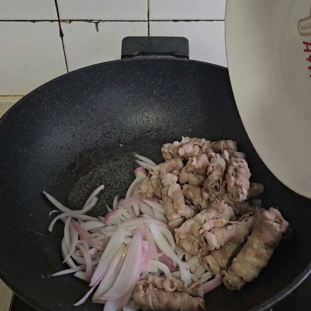 Blanchiertes Rindfleisch wird mit angebratenen Zwiebeln in einer heißen Pfanne vermengt.
