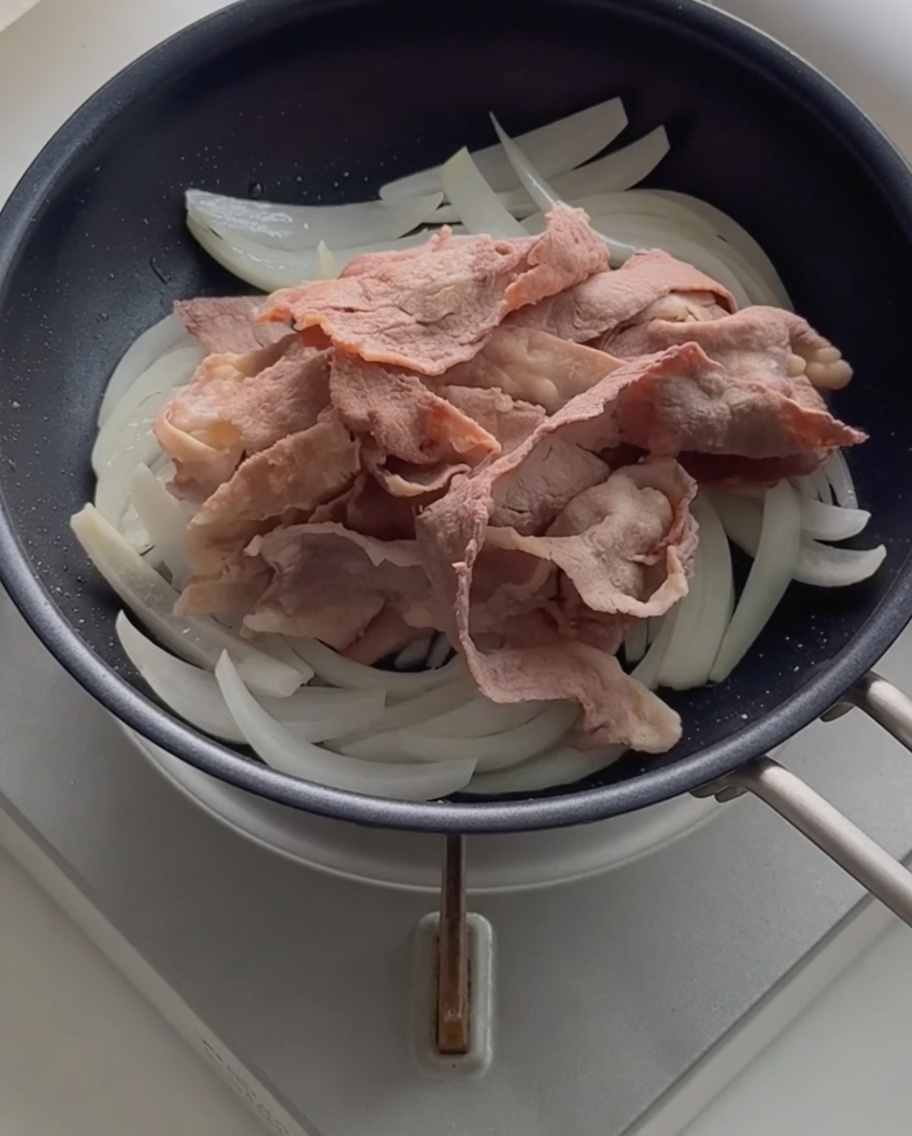 Blanchiertes Rindfleisch liegt auf den angeschwitzten Zwiebeln in der Pfanne.