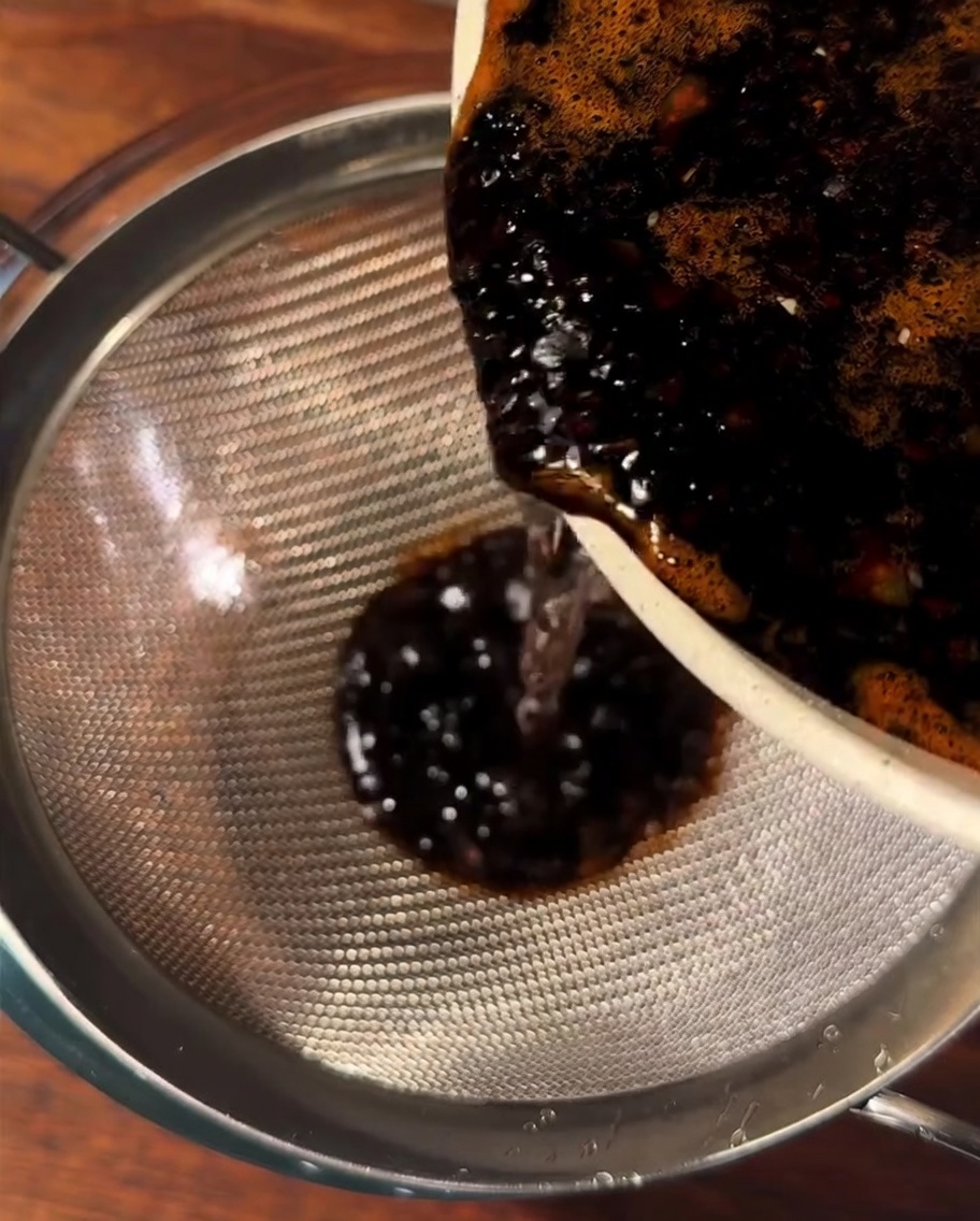 Eine dunkelbraune Sauce wird durch ein feines Metallsieb in eine Glasschüssel gegossen.