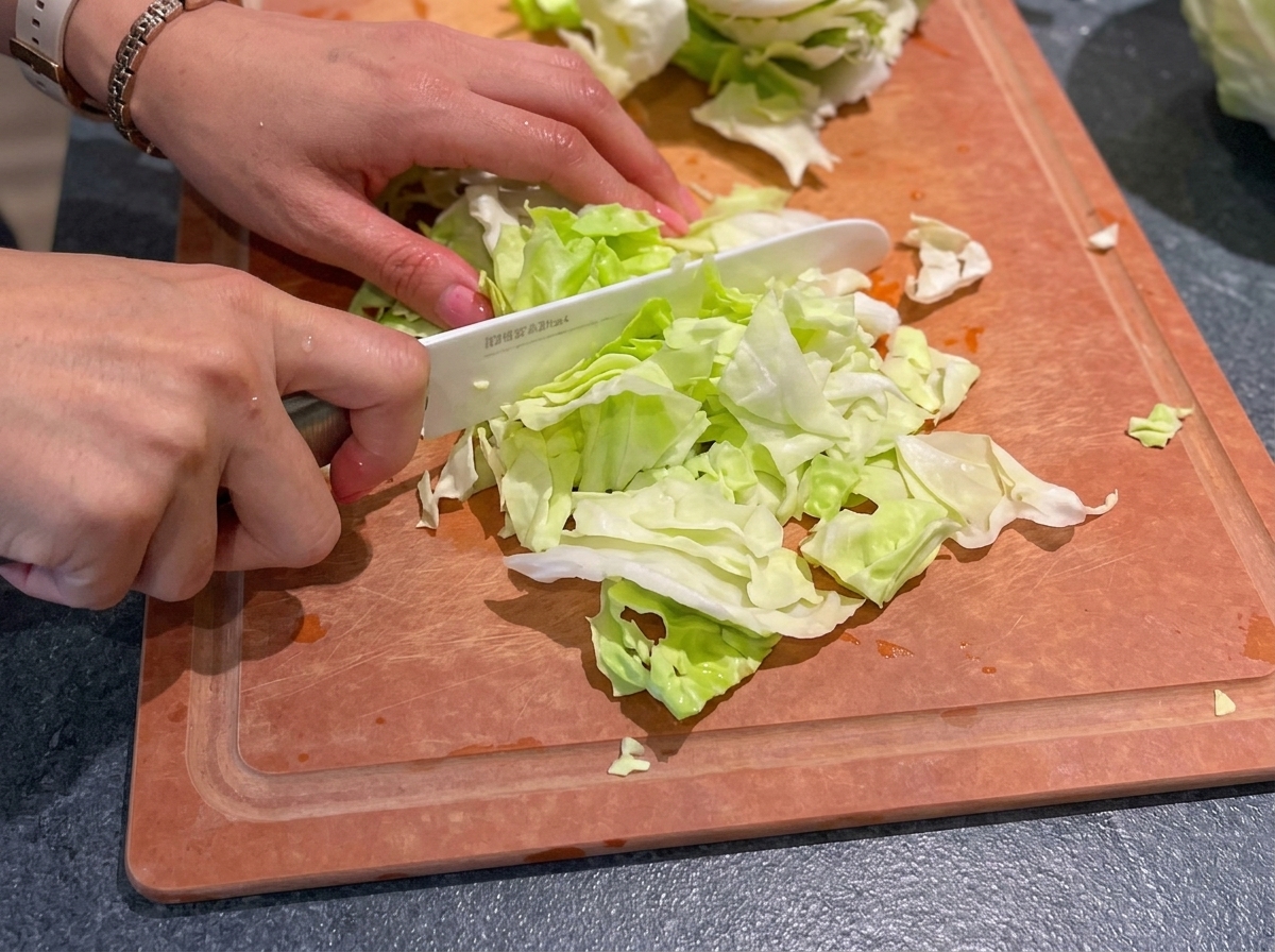 Hände schneiden mit einem weißen Keramikmesser frischen grünen Weißkohl auf einem Holzschneidebrett in kleine Stücke.