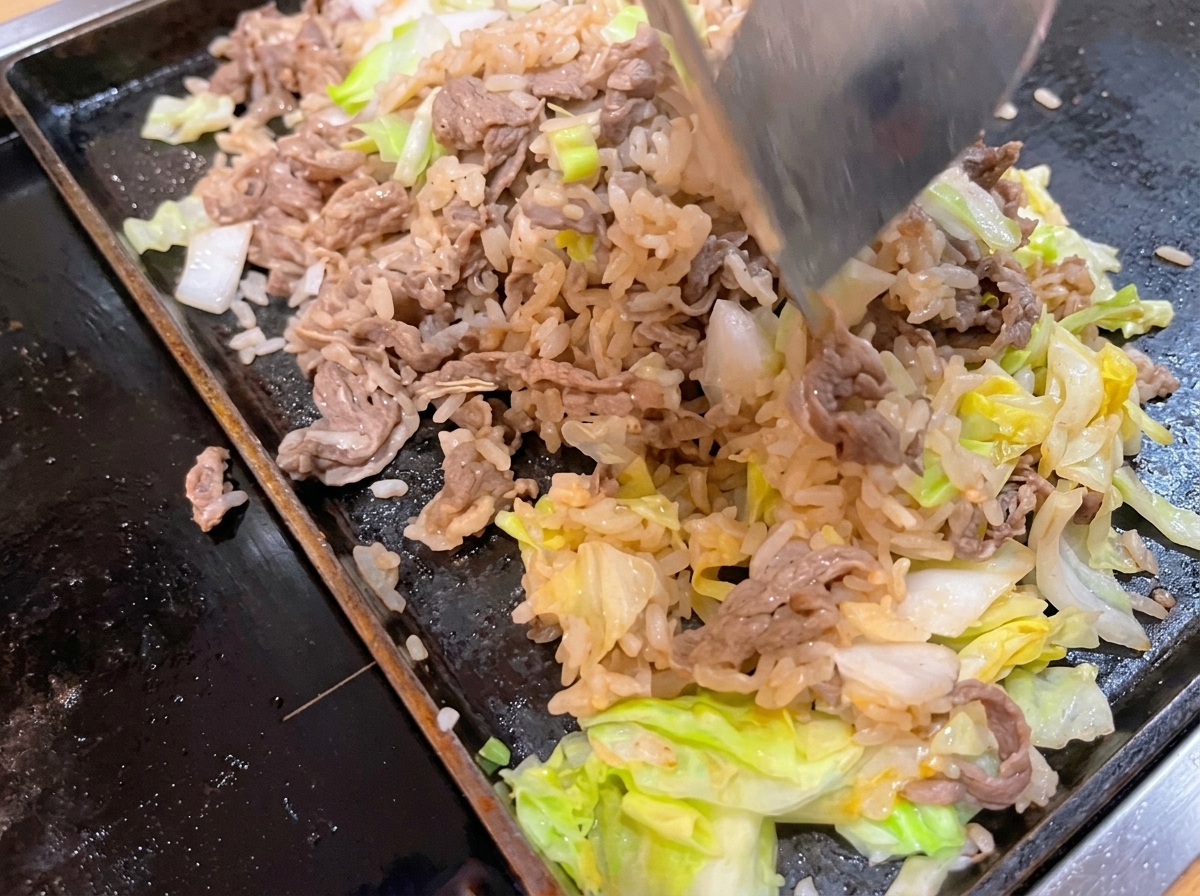 Ein Metallspatel mischt gebratenes Rindfleisch, weißen Reis und grünen Kohl in einer heißen flachen Pfanne zusammen.
