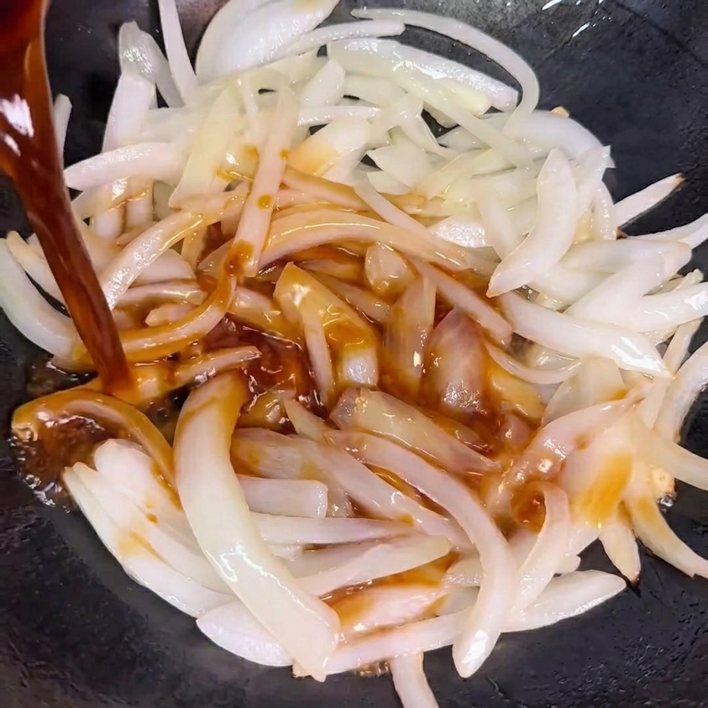 Die Sauce wird über die gebratenen Zwiebeln im Wok gegossen.