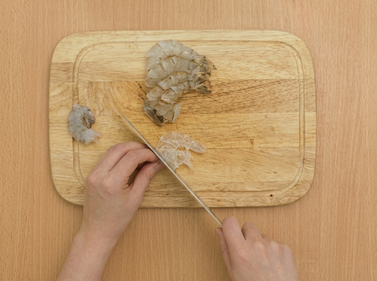 Nahaufnahme eines Messers, das flache Schnitte entlang des Bauchs einer rohen Garnele auf einem Holzbrett macht.