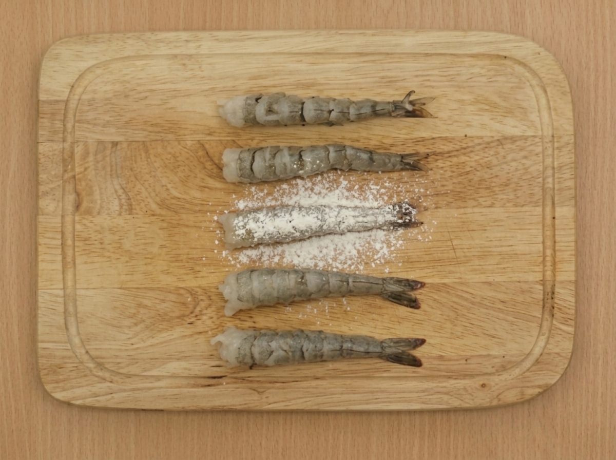 Ein feines Sieb streut Mehl über mehrere vorbereitete Garnelen auf einem Holzbrett.