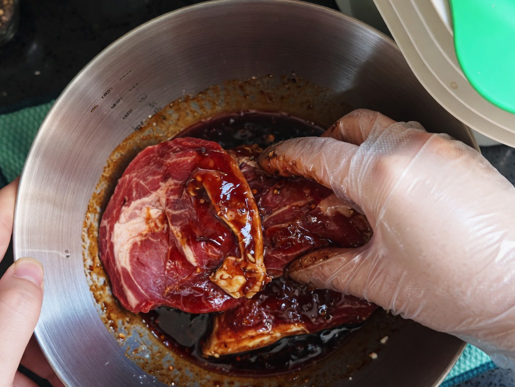 Eine Hand in einem Plastikhandschuh vermengt rohe Schweinekoteletts mit der Marinade in einer Metallschüssel.