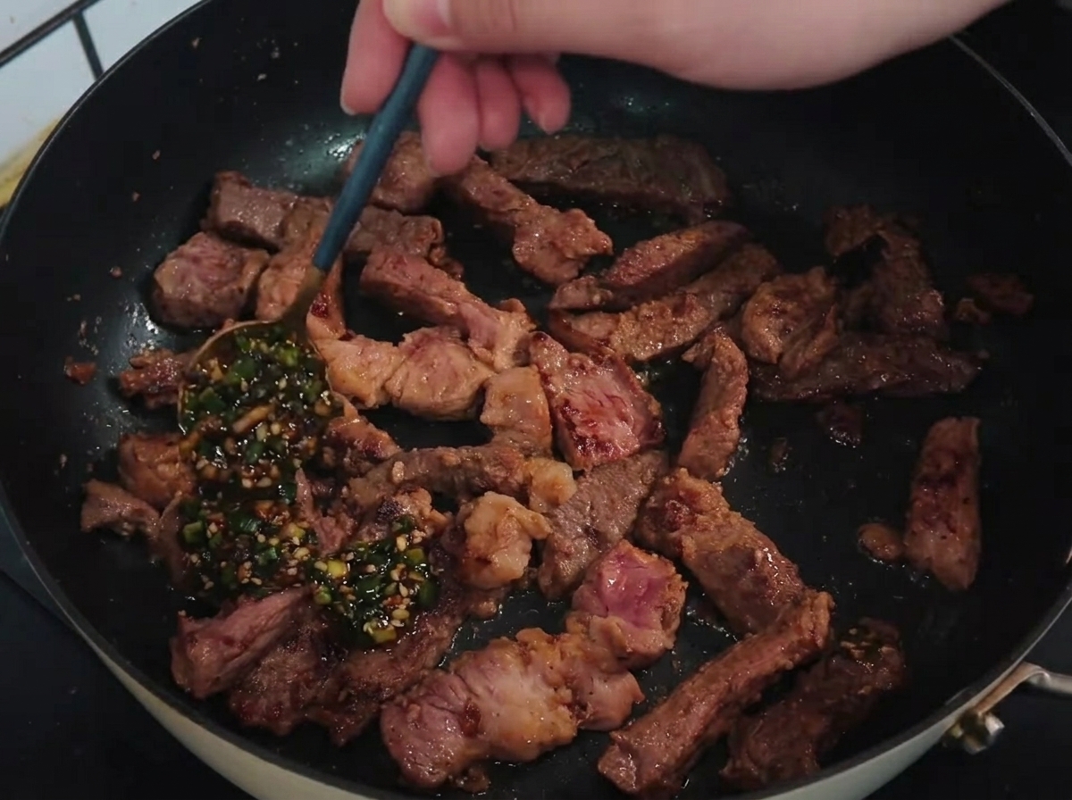 Ein Löffel gießt eine leuchtend grüne Frühlingszwiebel-Knoblauch-Sauce über angebratene Kotelettstreifen in einer heißen Pfanne.