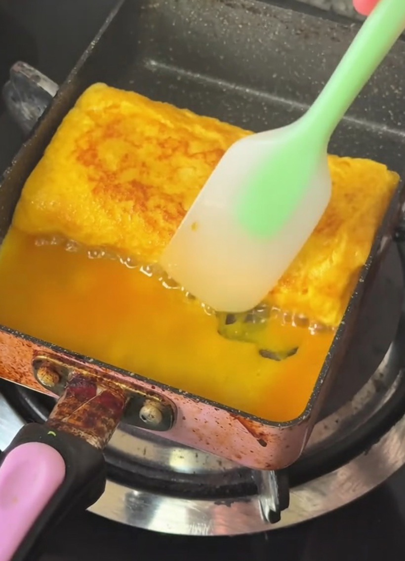 Eine rechteckige Pfanne mit einem geschichteten, goldgelben Omelett, das mit einem Silikonspatel gefaltet wird.