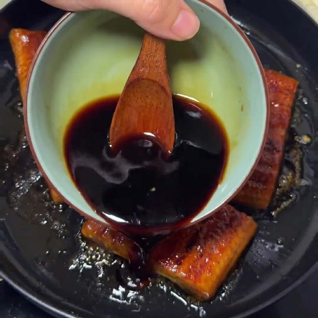Eine dunkle, würzige Sauce wird aus einer kleinen Schüssel über goldbraune Aalfilets in einer Pfanne gegossen.