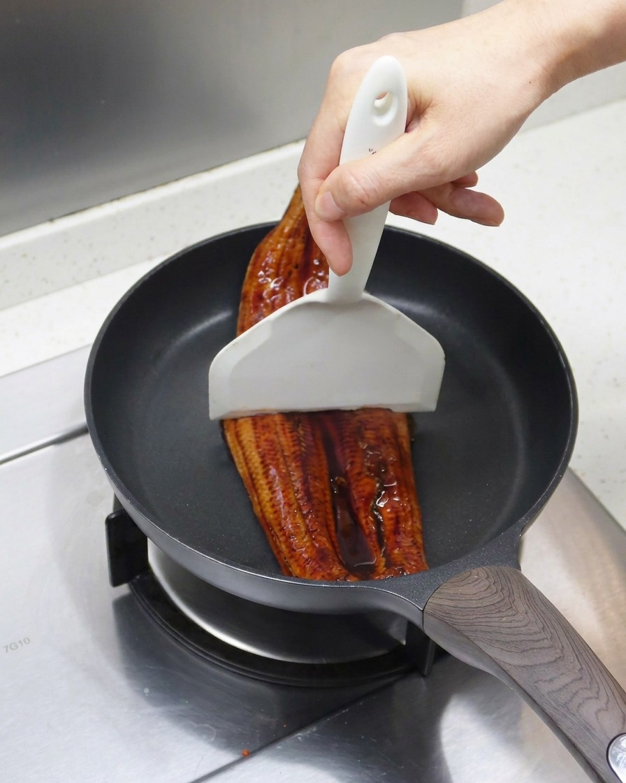 Der gegrillte Aal wird mit einem weißen Pfannenwender in der Pfanne in zwei Stücke geteilt.