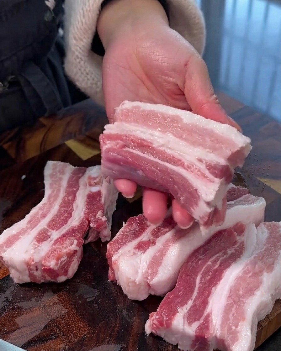 Roher Schweinebauch wird auf einem Holzschneidebrett in gleichmäßige, dicke Streifen geschnitten.