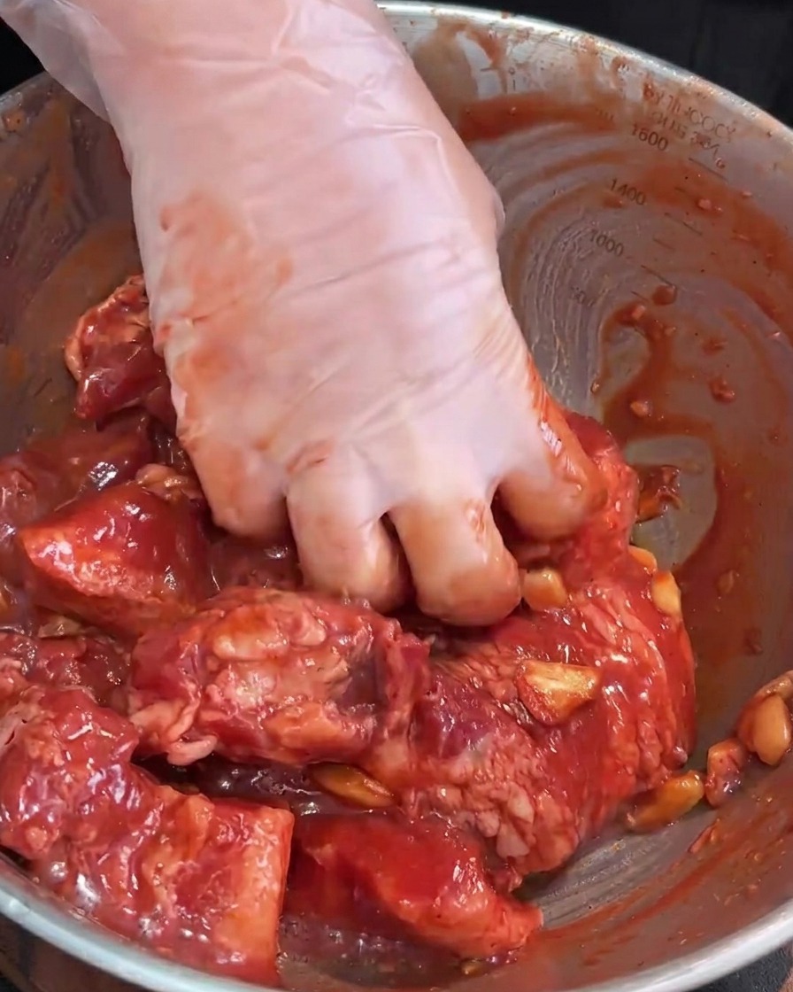 Behandschuhte Hände massieren die Marinade in den rohen Schweinebauch in einer Edelstahlschüssel.