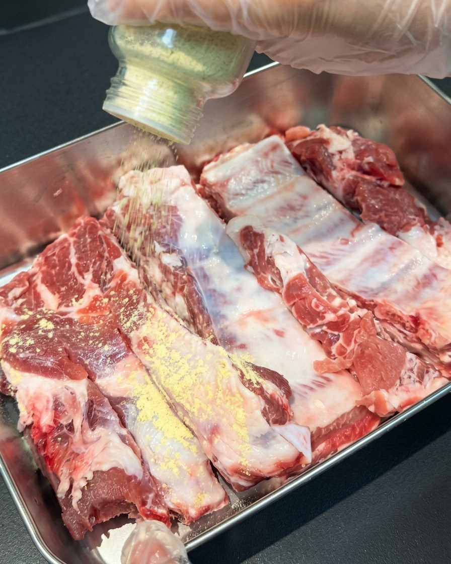 Eine Hand streut gelbes Knoblauch- und Ingwerpulver aus einem Glasstreuer auf rohe Schweinerippchen in einer Metallschale.