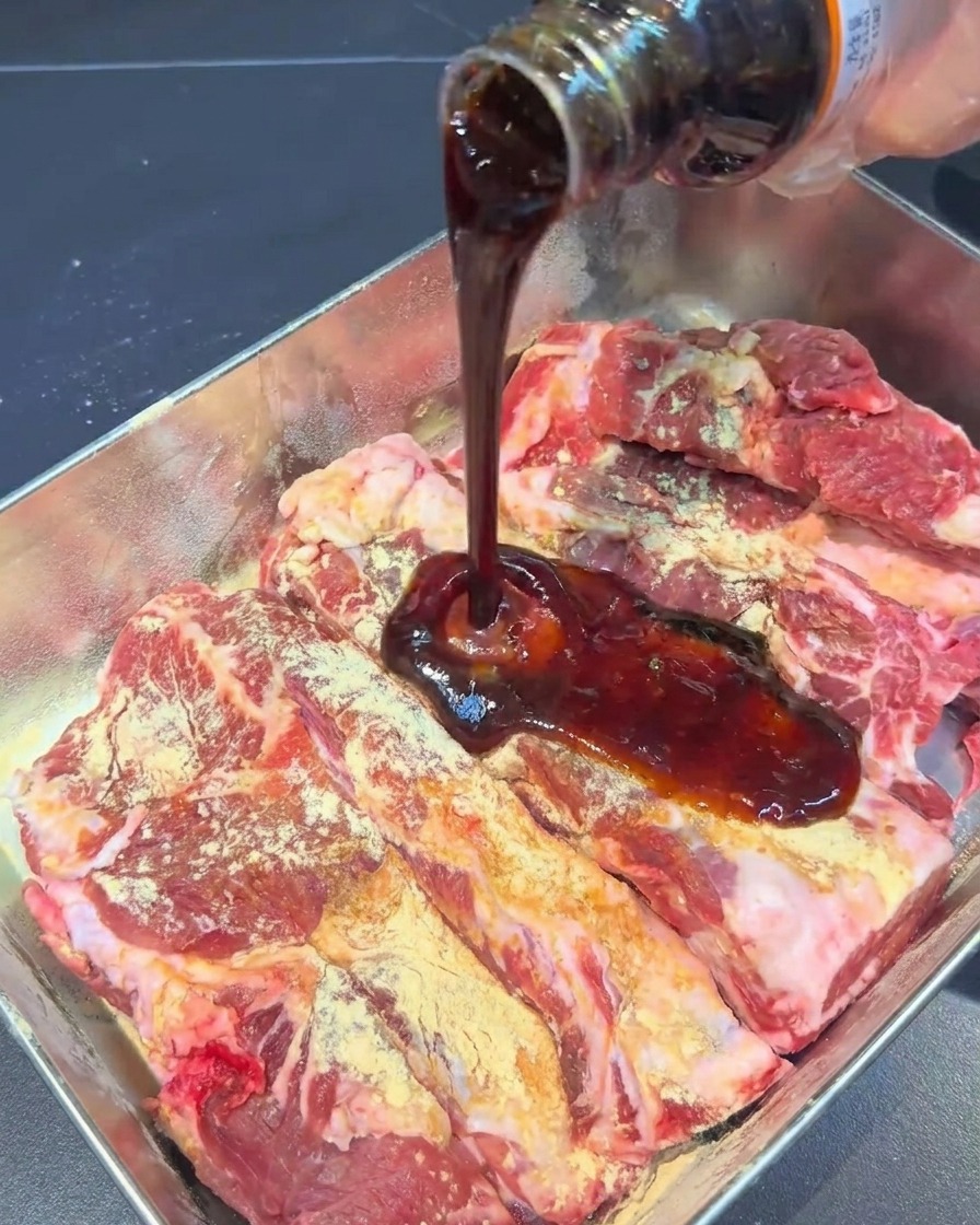 Dunkle Marinadensauce wird aus einer Glasflasche auf gewürzte Schweinerippchen in einer Metallschale gegossen.