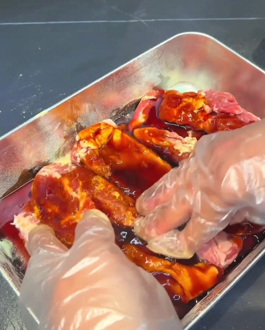 Schweinerippchen werden mit behandschuhten Händen in einer dunklen Marinadensauce in einem Metallbehälter massiert und bestrichen.