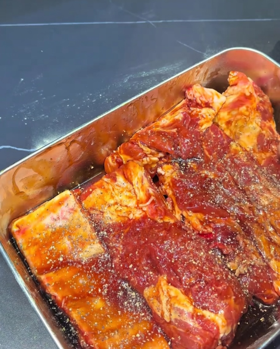 Nahaufnahme von rohen Schweinerippchen, die komplett mit einer dunklen Marinade überzogen und mit grobem schwarzen Pfeffer bestreut sind.
