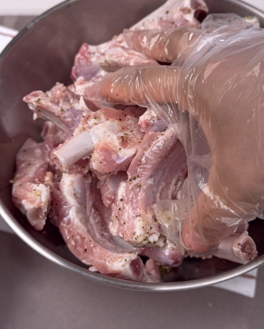 Hände mit Plastikhandschuhen mischen die gewürzten Schweinerippchen in einer Edelstahlschüssel.