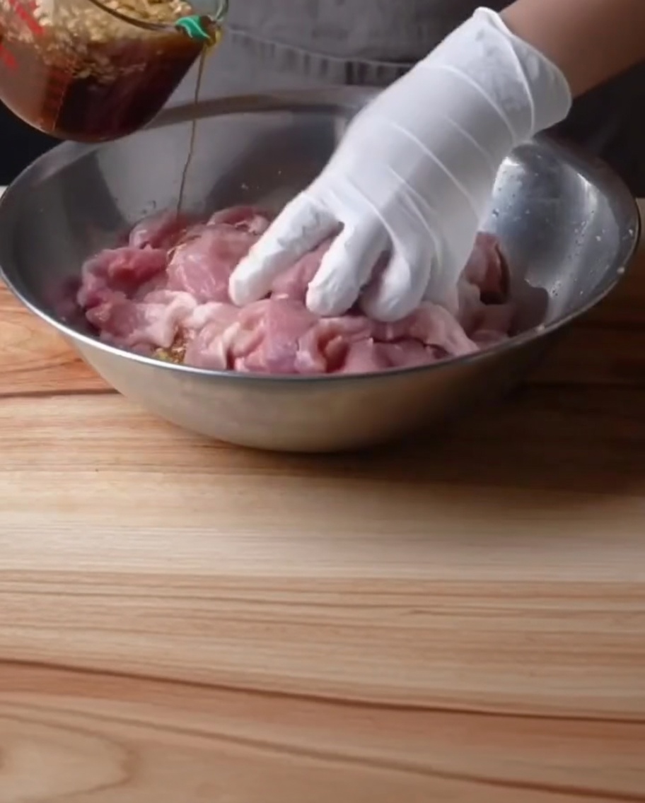 Eine behandschuhte Hand gießt dunkle Marinade über rohe Schweinerippchen in einer Metallschüssel.