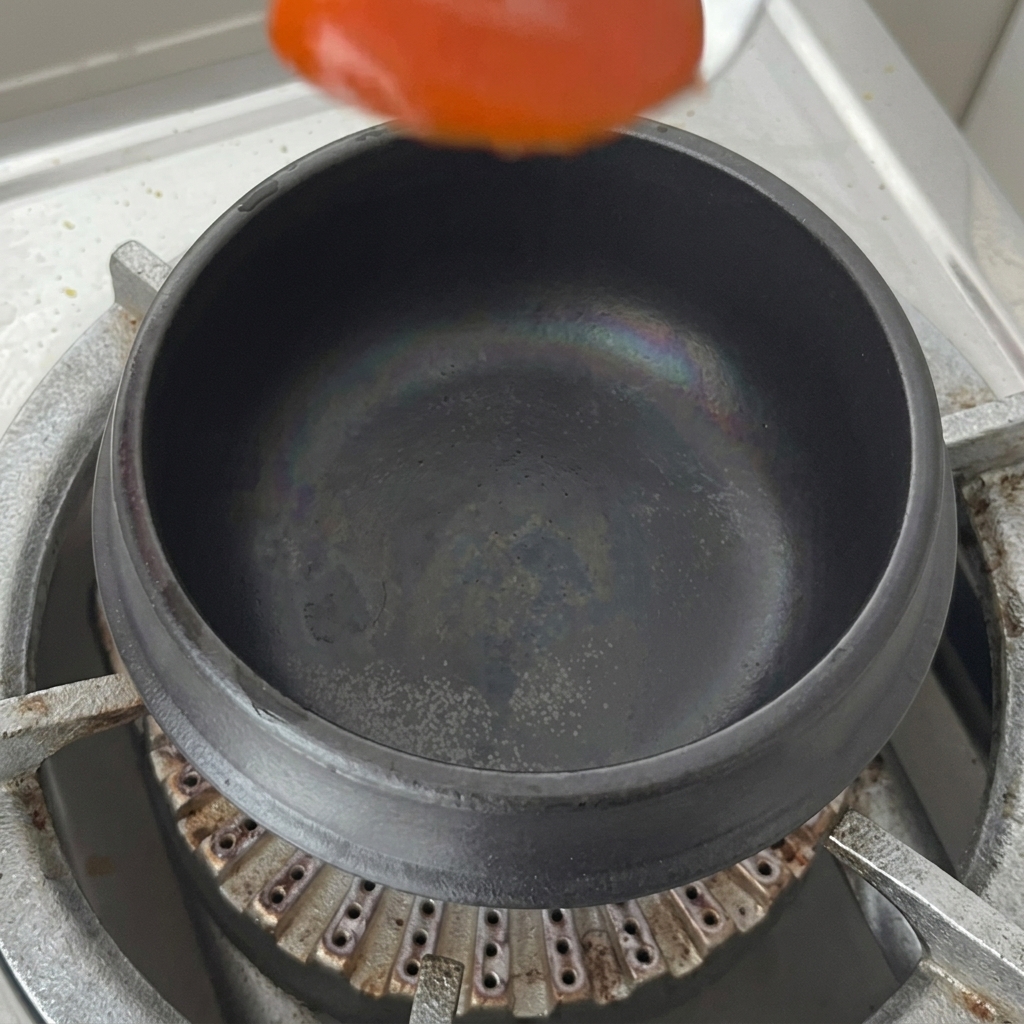 Ein Löffel rote Chilipaste wird in einen leeren schwarzen Tontopf auf dem Herd gegeben.