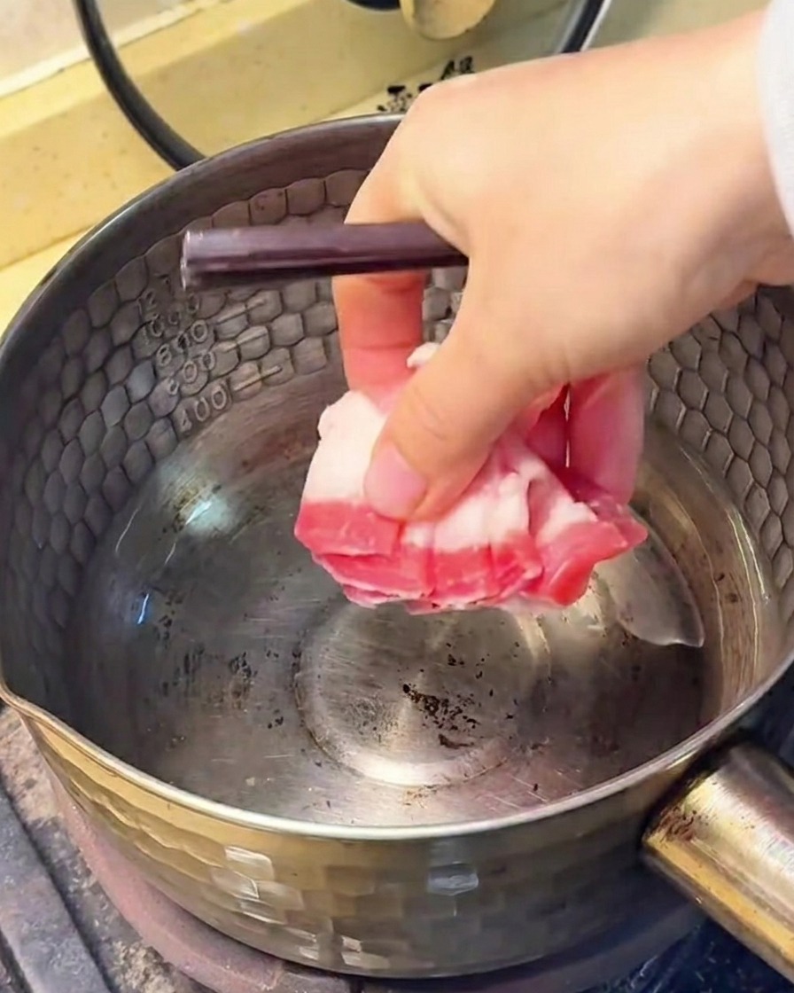 Schweinebauchscheiben braten in einem Edelstahltopf, wobei das ausgelassene Fett am Boden sichtbar ist.