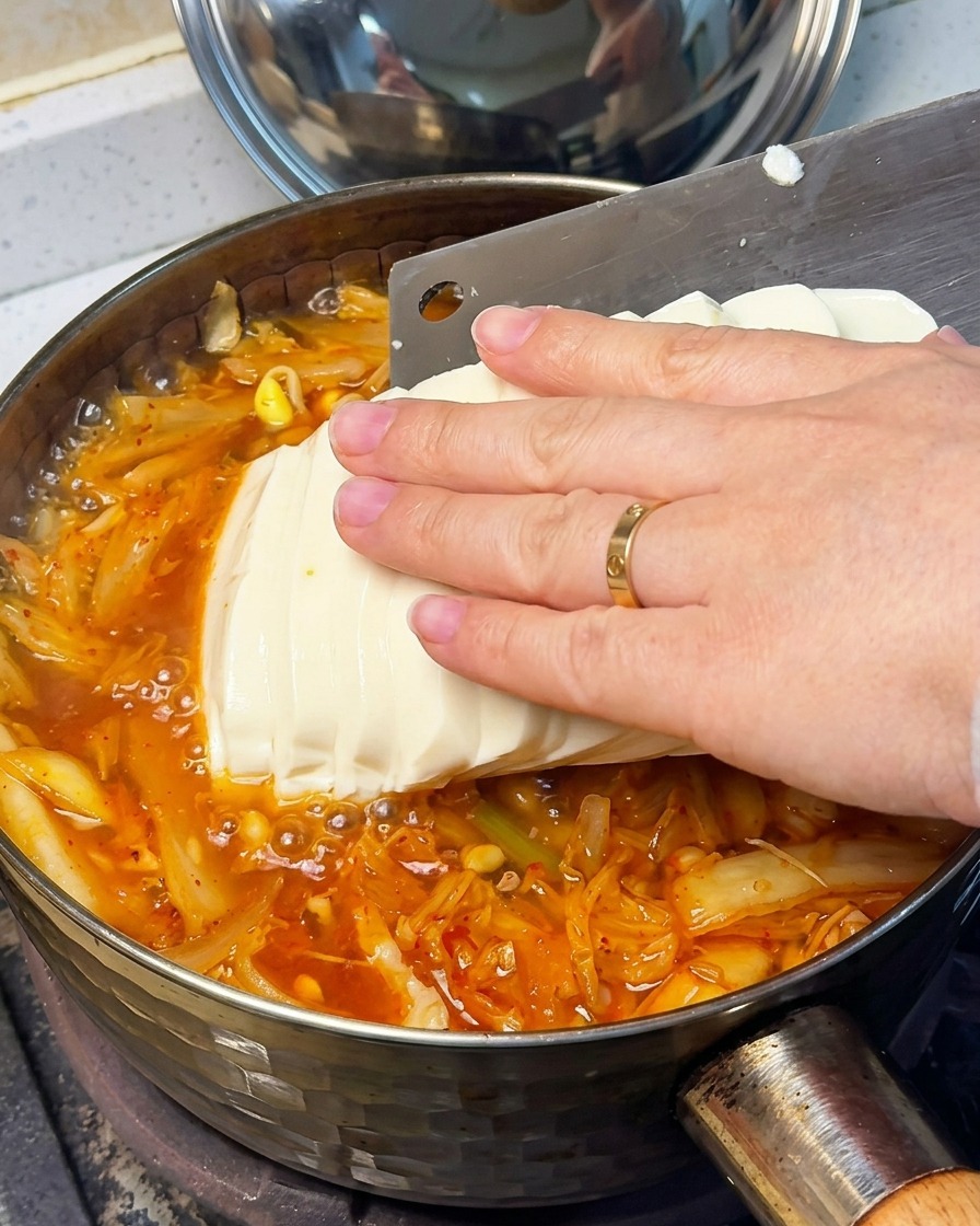 Nahaufnahme einer Hand, die vorsichtig geschnittenen Seidentofu in die sprudelnde rote Kimchi-Suppe gibt.