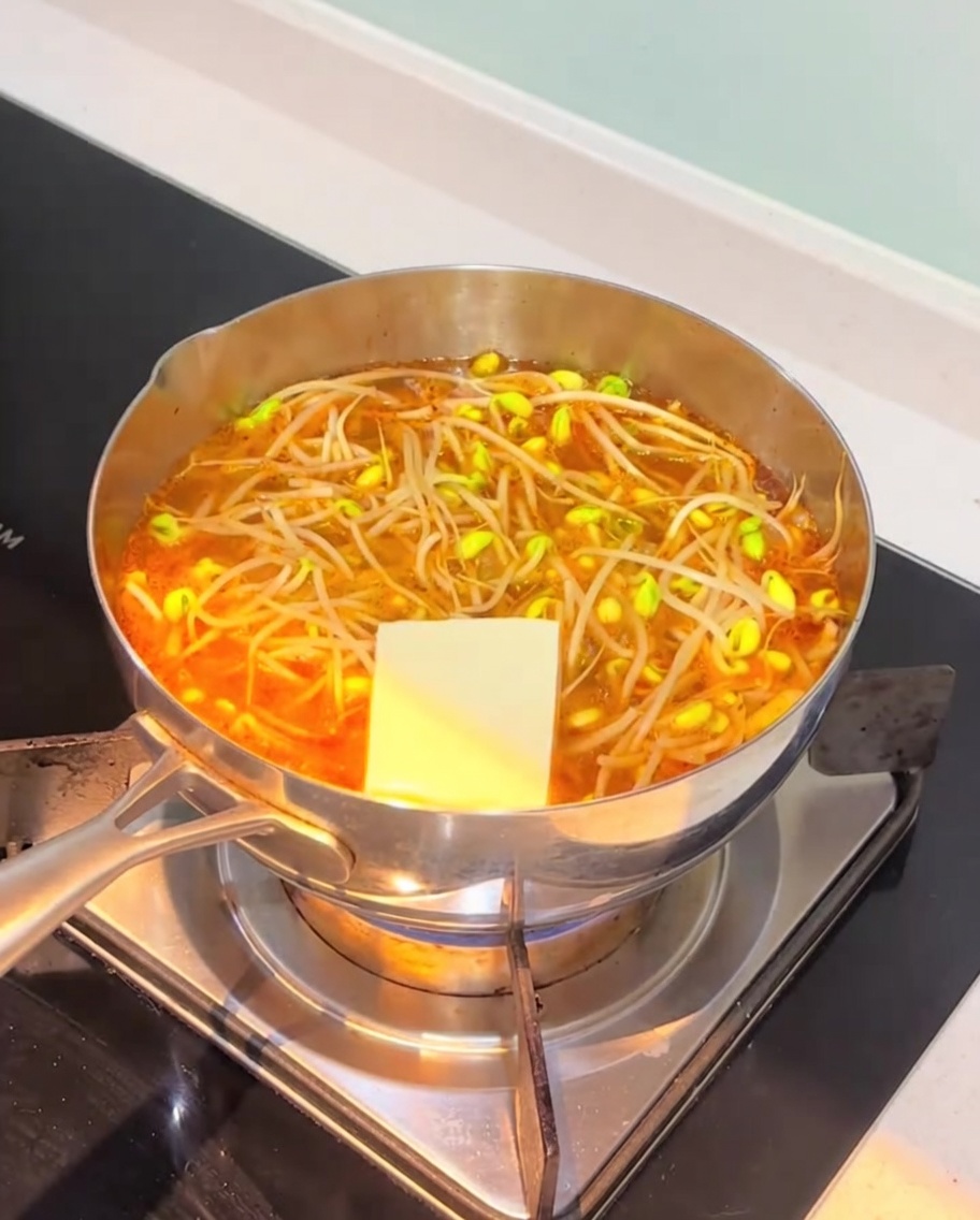 Scheiben von weißem Tofu und Fischkuchen werden in die köchelnde orangefarbene Kimchi-Suppenbasis gegeben.