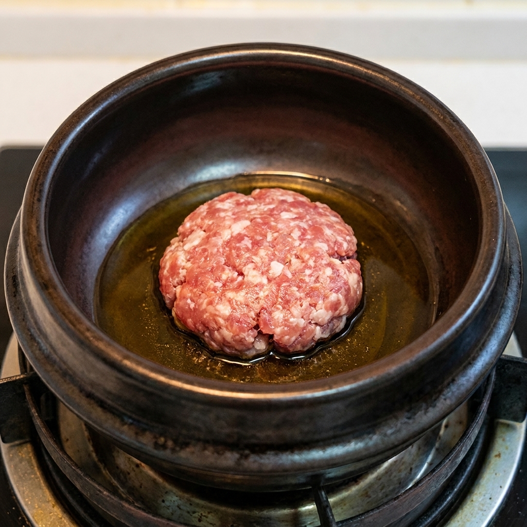 Eine Kugel Hackfleisch bräunt in heißem Sesamöl in einem dunklen Tontopf.