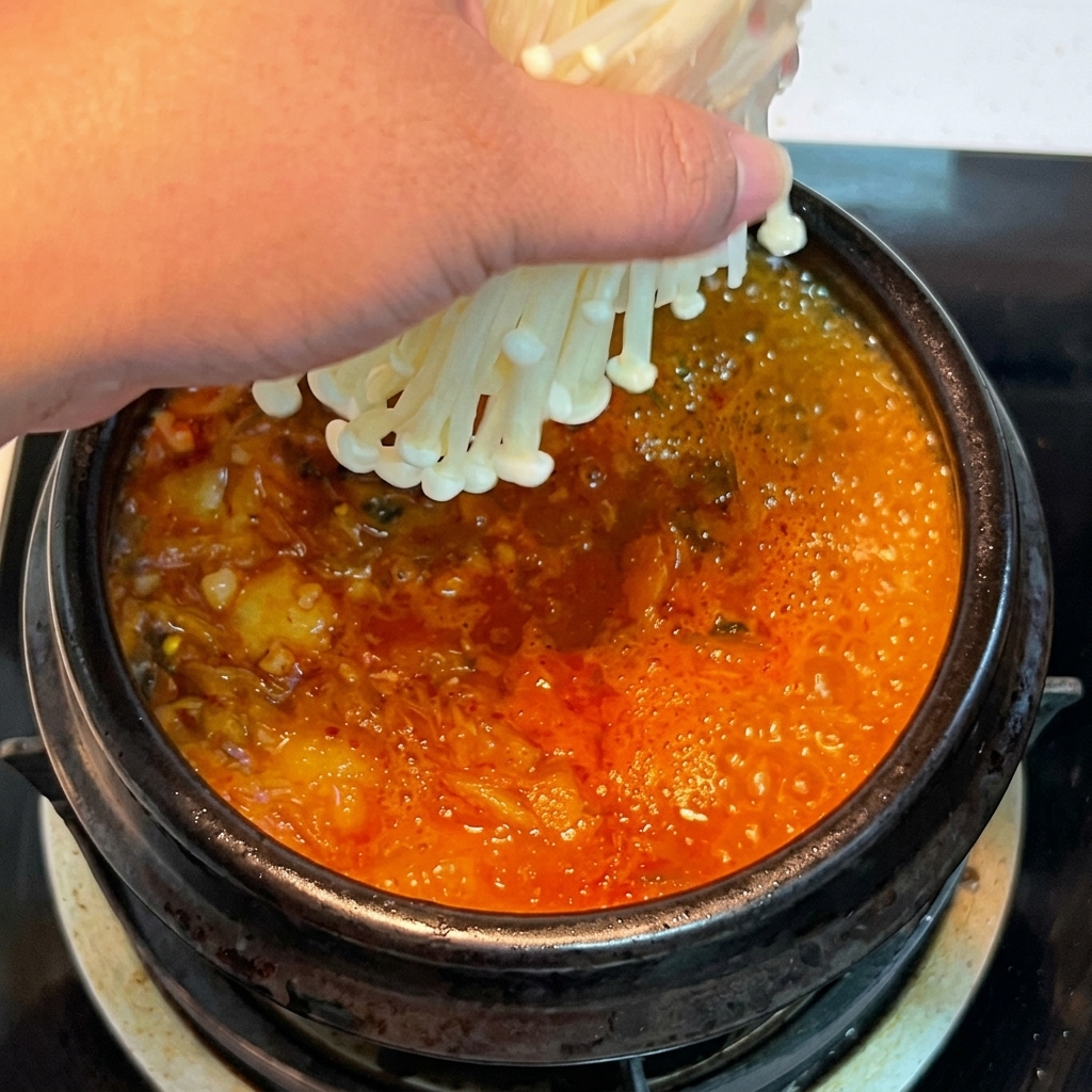 Eine Hand platziert ein Bündel frischer Enoki-Pilze in den sprudelnden, roten Kimchi-Eintopf.