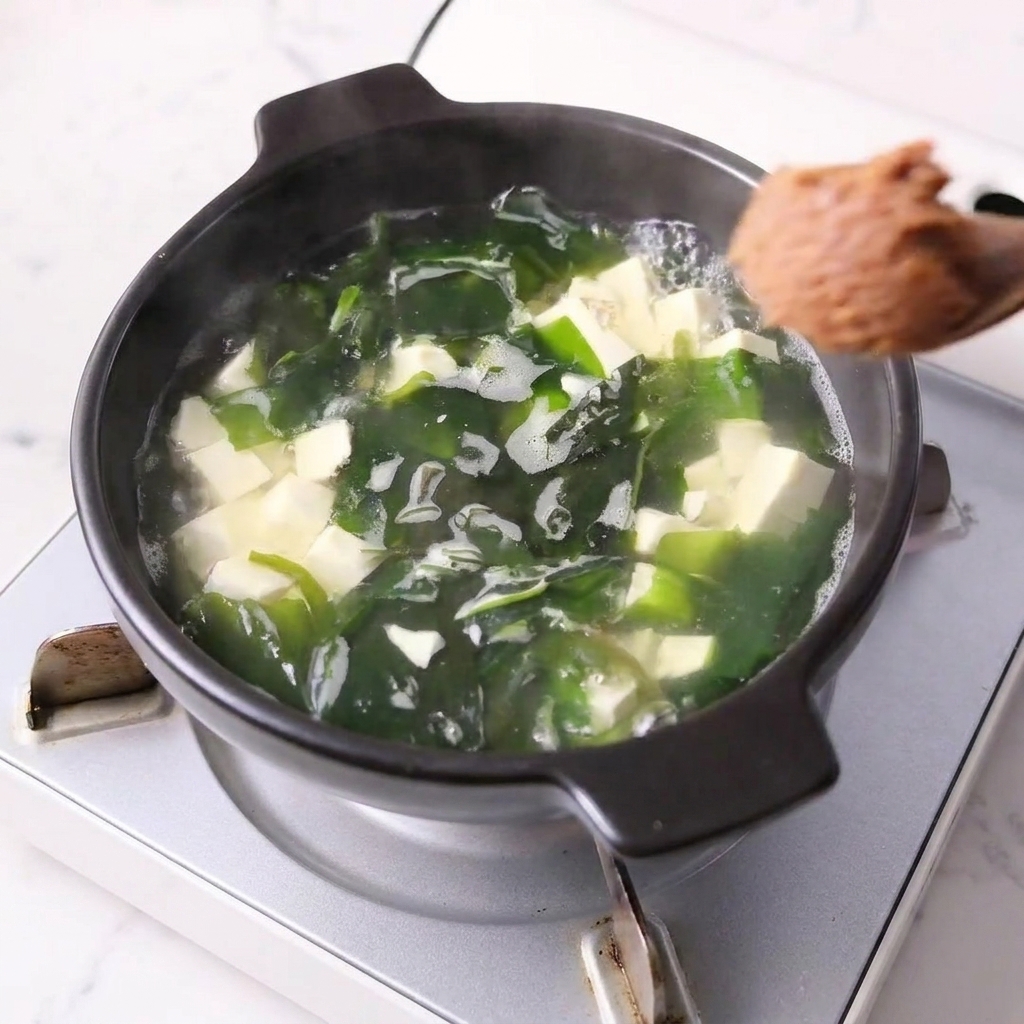 Ein Löffel Miso-Paste wird in einen Topf mit Tofu und Wakame gerührt.