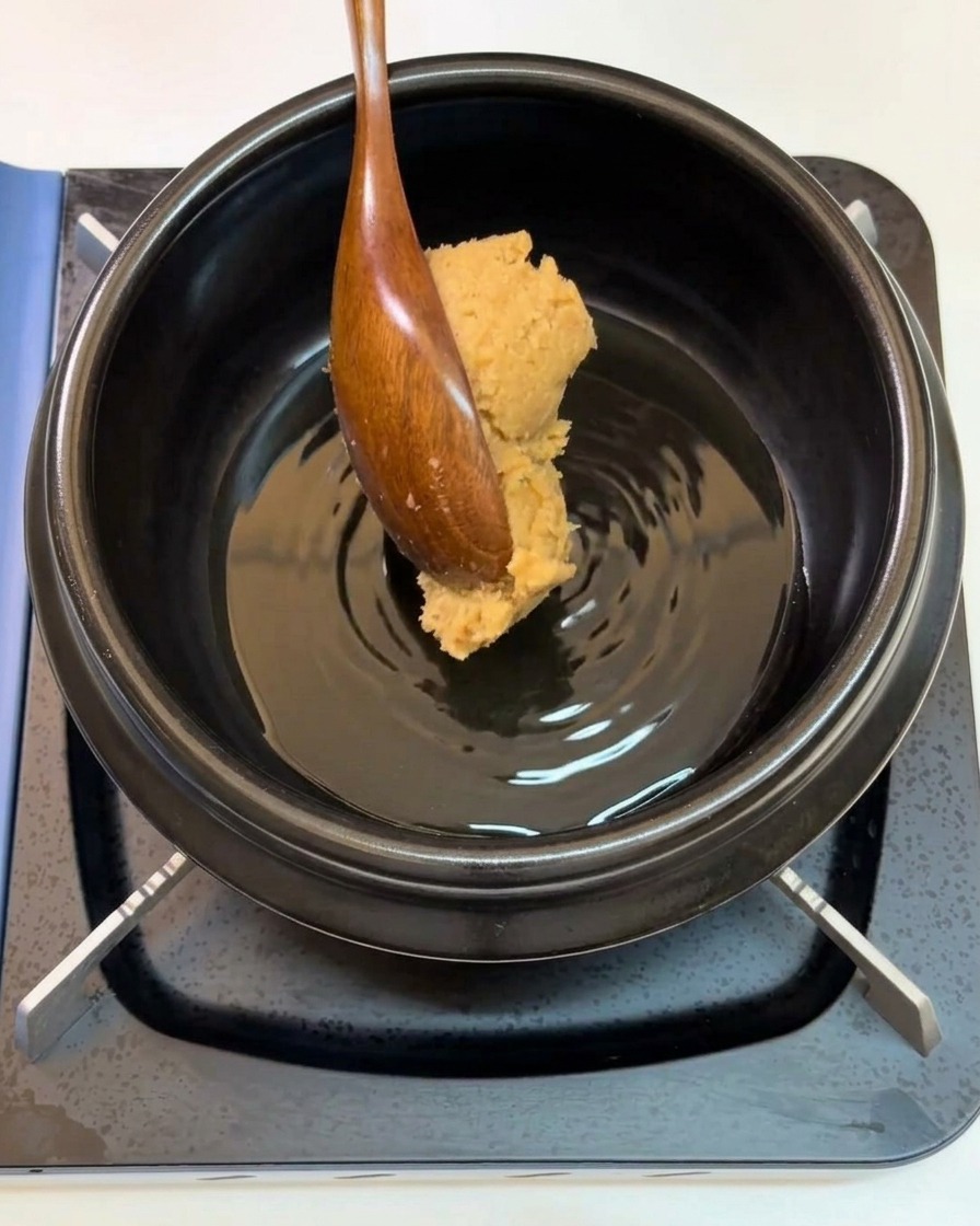 Ein Holzlöffel gibt einen Klecks Sojabohnenpaste in den Topf.